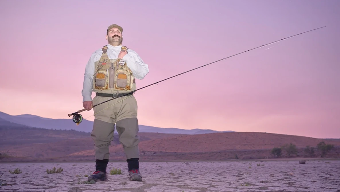 Fly fisherman in waders standing in shallow water at dusk with mountains in background.