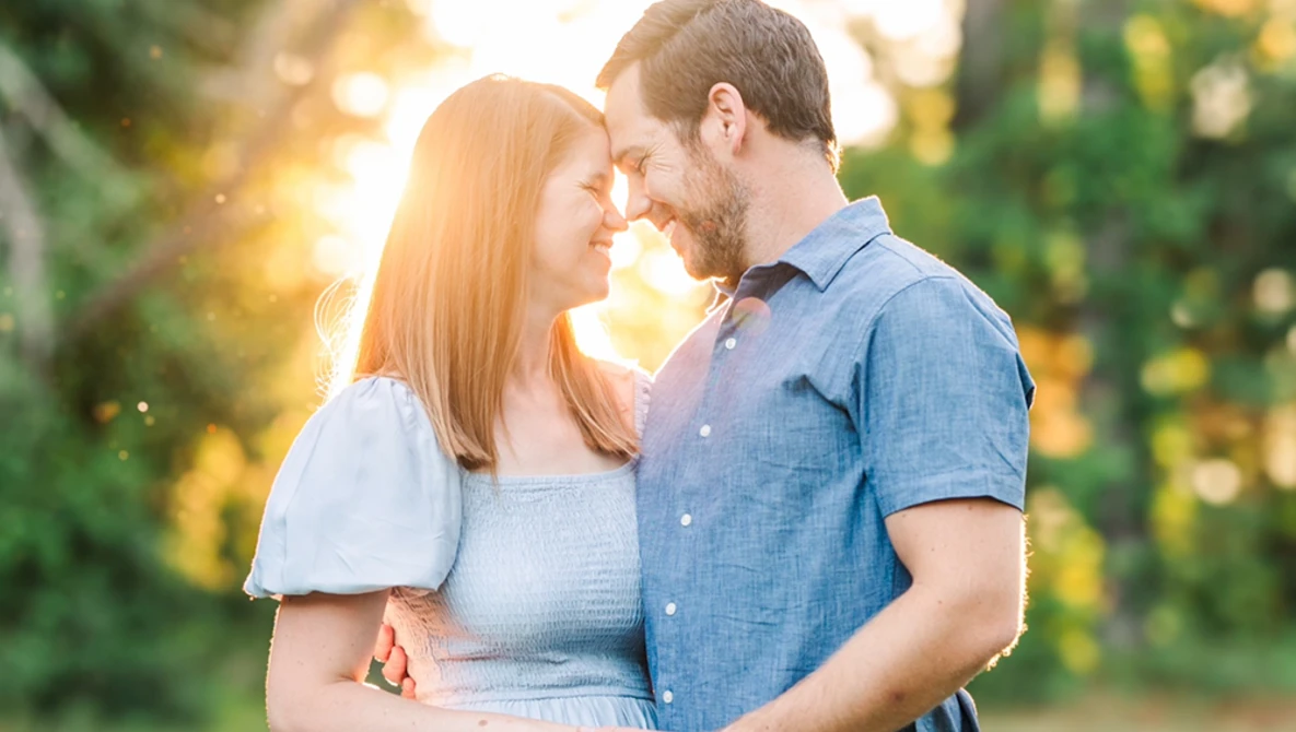 Couple embracing outdoors with foreheads touching, backlit by golden sunlight filtering through trees.