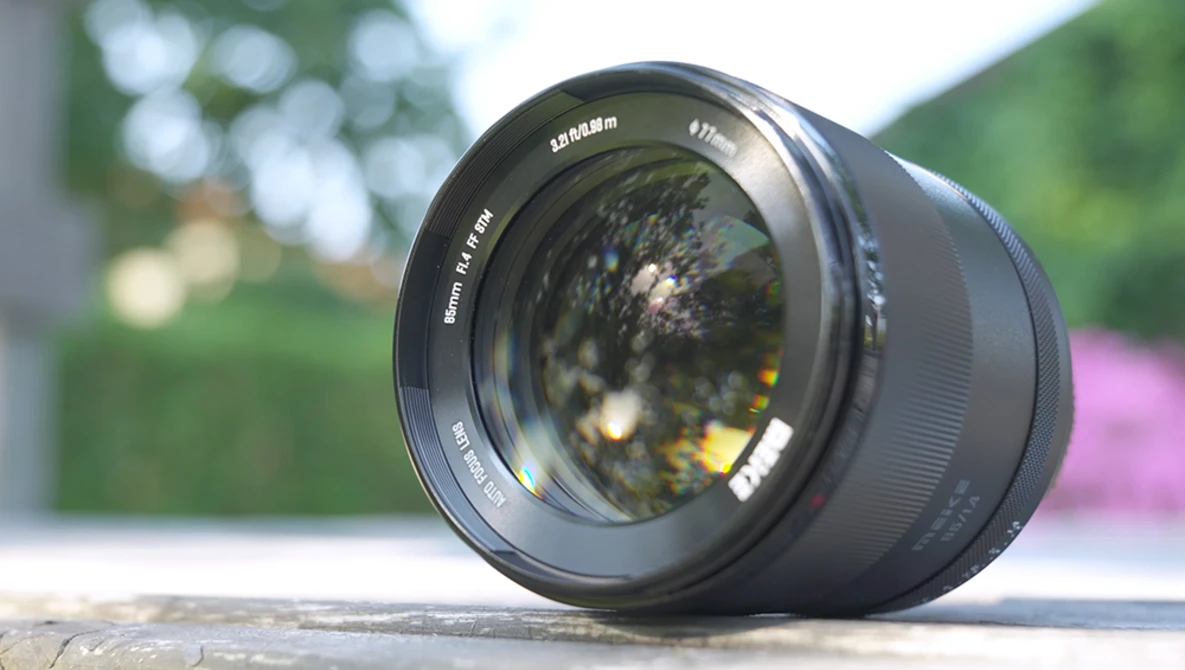 Black prime lens positioned on wooden surface with blurred garden and flowering plants in background.
