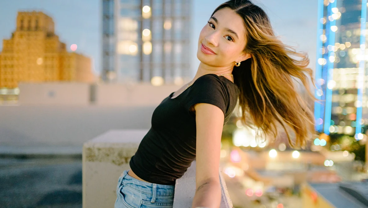 Woman in black crop top and jeans posing on rooftop at dusk with city skyline behind her.