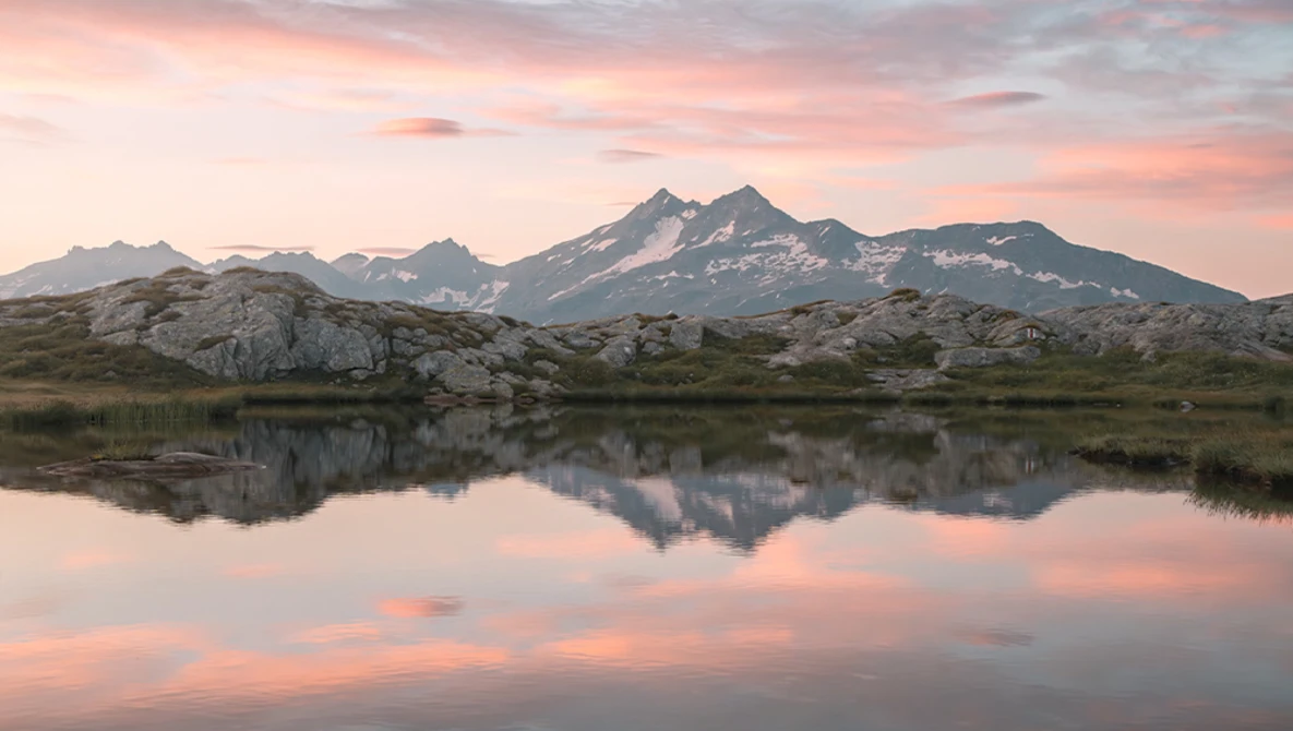 Snow-capped mountain reflected in still lake water at sunset with pink and orange sky.