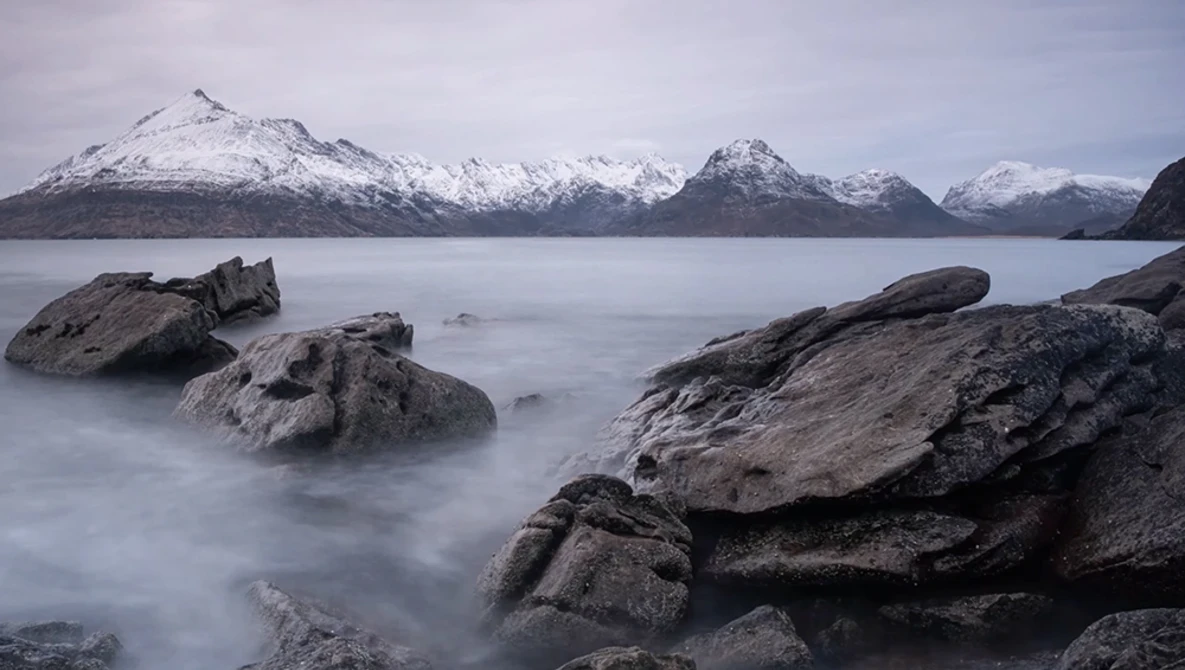 Snow-capped mountains across a calm fjord with foreground rocks smoothed by long-exposure water effect.