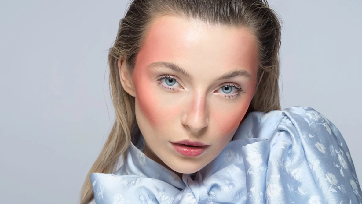 Portrait of a woman with blue eyes and pink-toned cheeks against a neutral gray background.