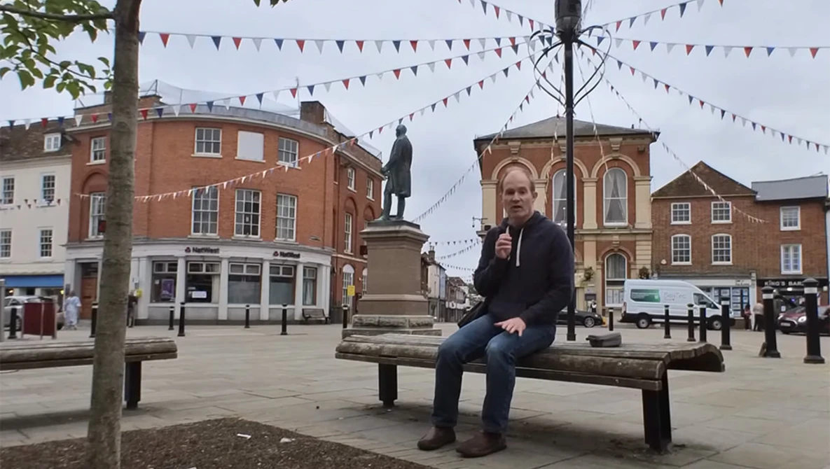 Person sitting on a wooden bench in a town square surrounded by Victorian-era brick buildings and festive bunting.