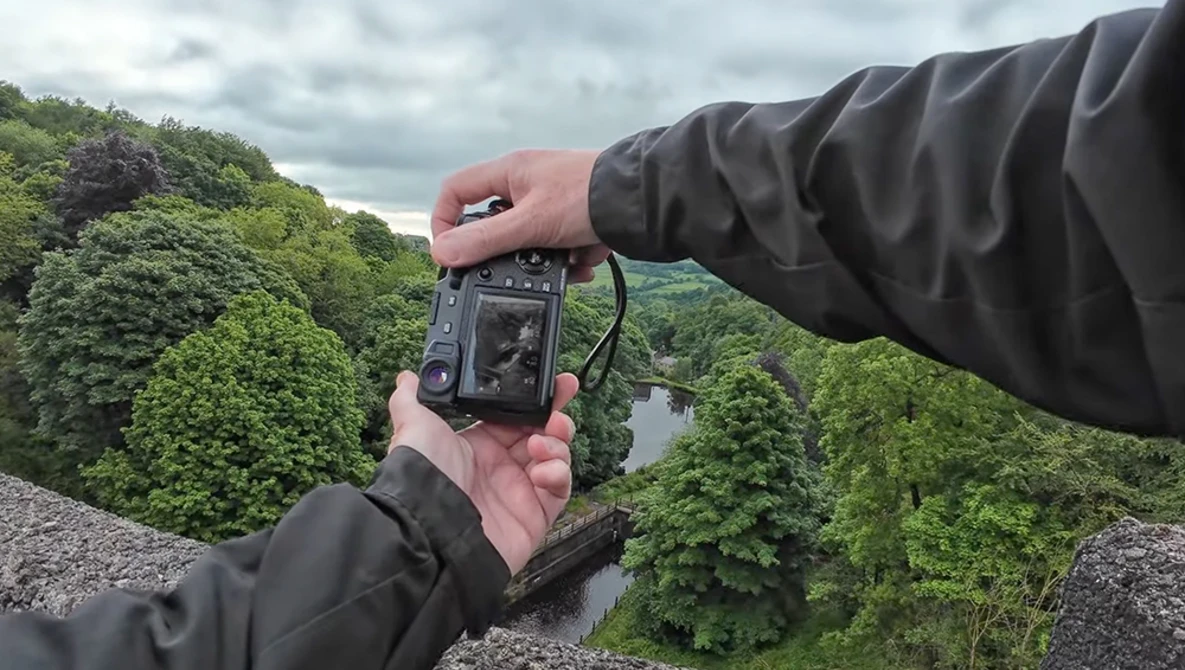Hands holding a thermal imaging camera pointed at a lush green valley landscape with a river visible below.
