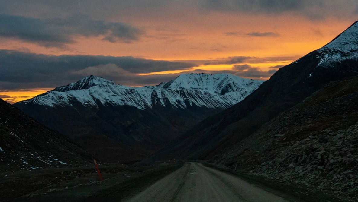 Straight gravel road stretching toward snow-capped mountains under a golden sunset sky.
