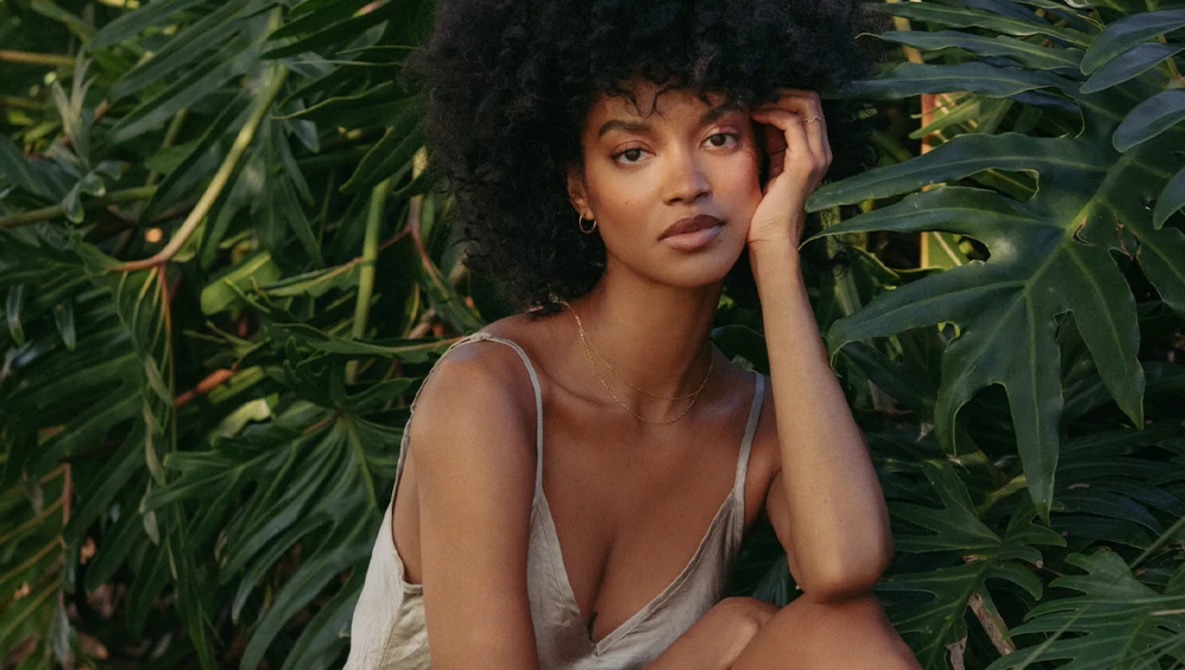 Woman with voluminous natural hair wearing a cream-colored top, posed among lush green foliage.