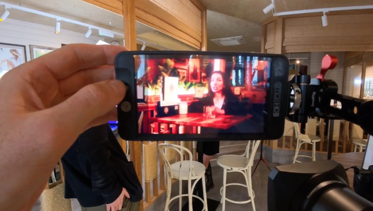 Hand holding smartphone displaying video content in a studio setting with red lighting and seated figures.
