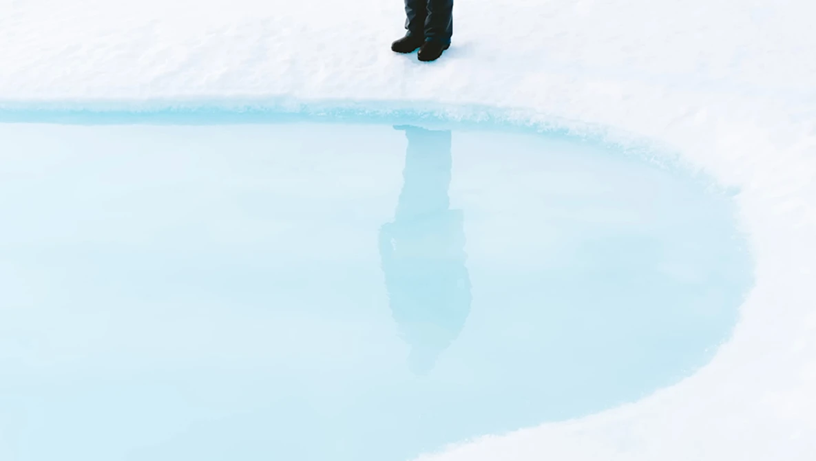 Person standing at the edge of a bright turquoise geothermal hot spring pool surrounded by white mineral deposits and snow.
