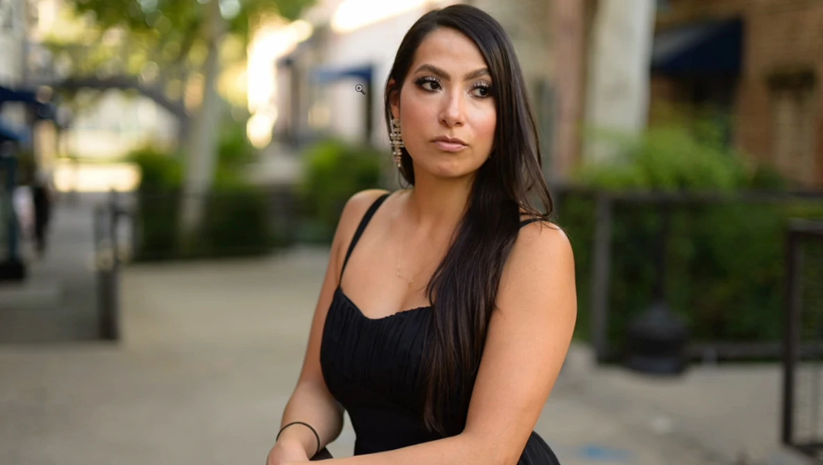 Woman in black sleeveless dress posing outdoors on urban street with blurred background.