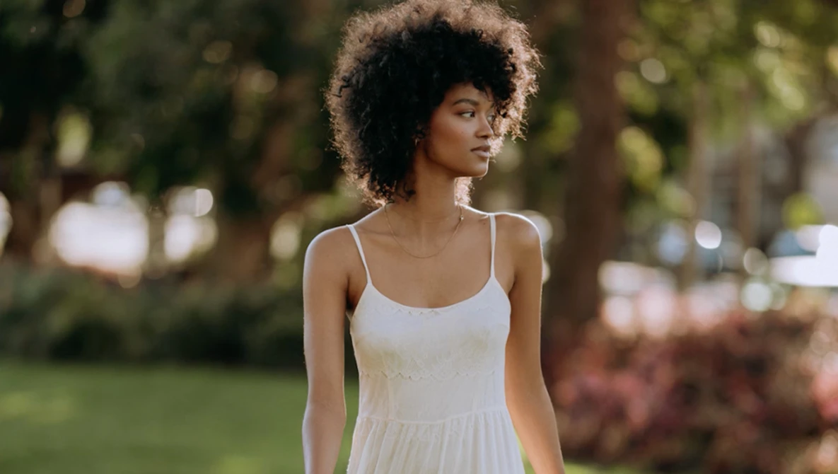 Woman in white camisole dress posing outdoors among trees and grass.