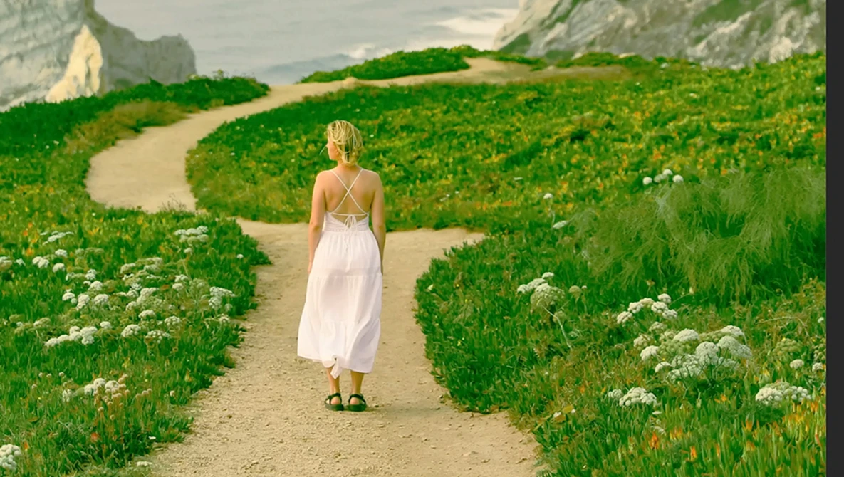 Woman in white dress walking down a dirt path through rolling green fields with white wildflowers.