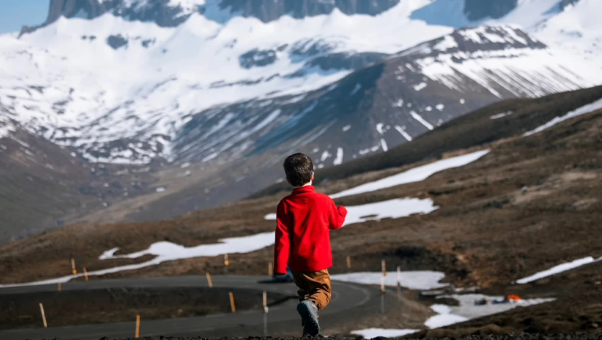 Runner in red jacket jogging on mountain road with snow-capped peaks and patches of snow in background.