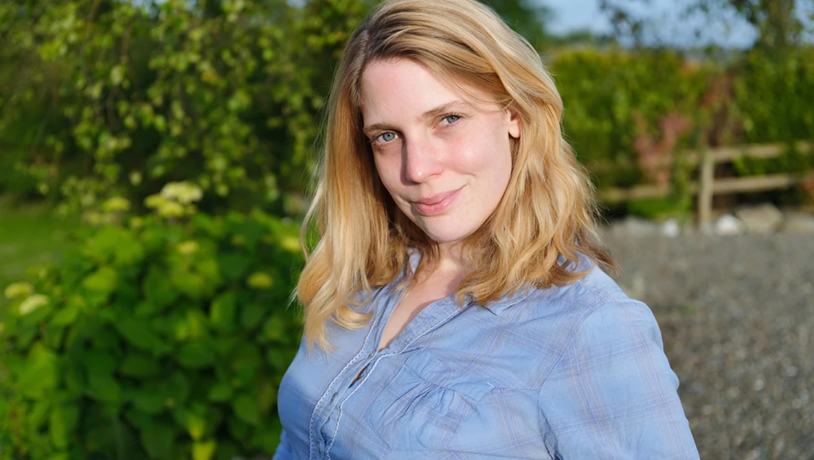 Woman with blonde hair wearing a light blue button-up shirt, photographed outdoors with green foliage in the background.