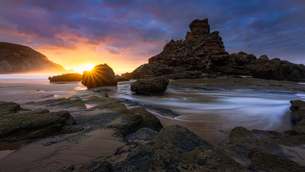 Rocky coastline at sunset with golden light between layered stone formations and smooth water in foreground.