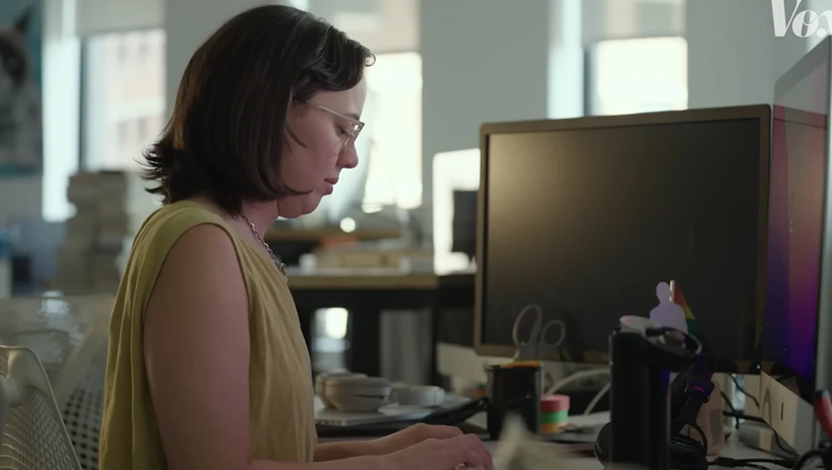 Woman in yellow sleeveless top working at computer in bright office space with large monitor.