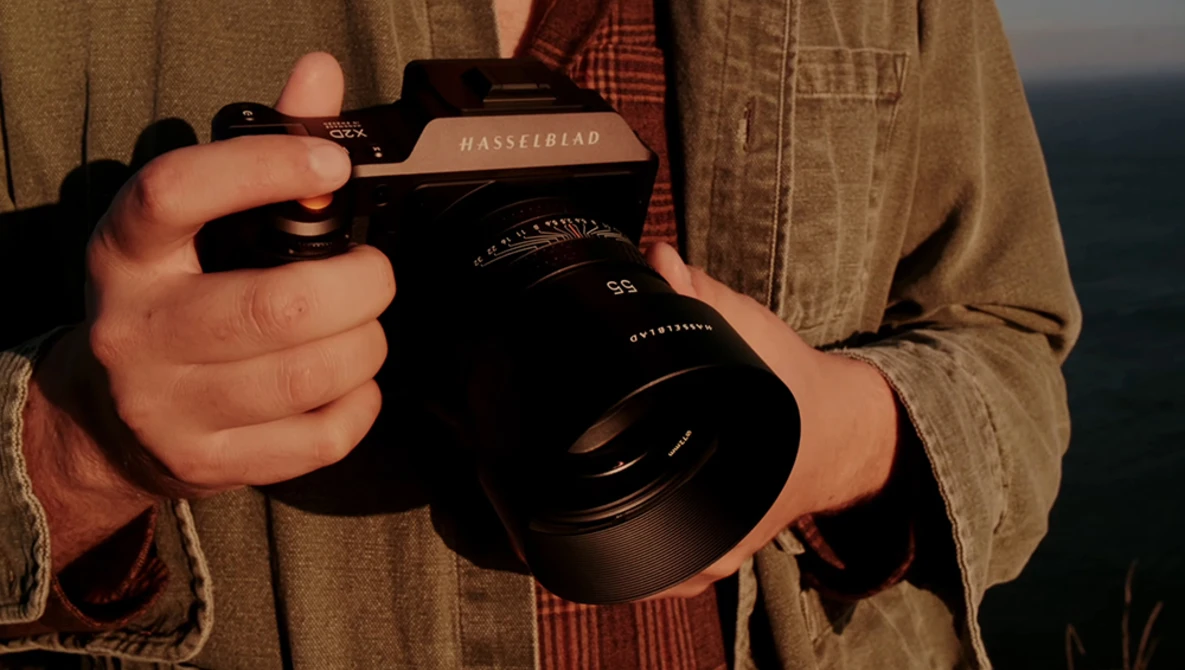 Photographer holding a Hasselblad camera with a large lens, wearing a tan button-up shirt.