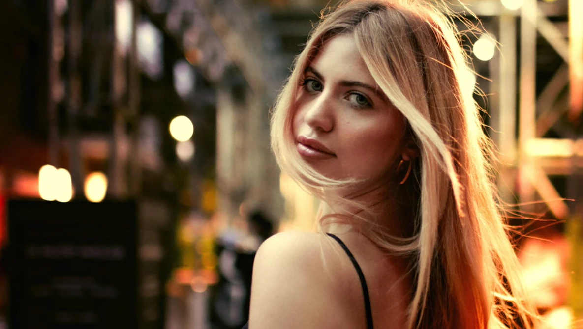Woman with long reddish-blonde hair looking over shoulder in urban evening setting with warm city lights.