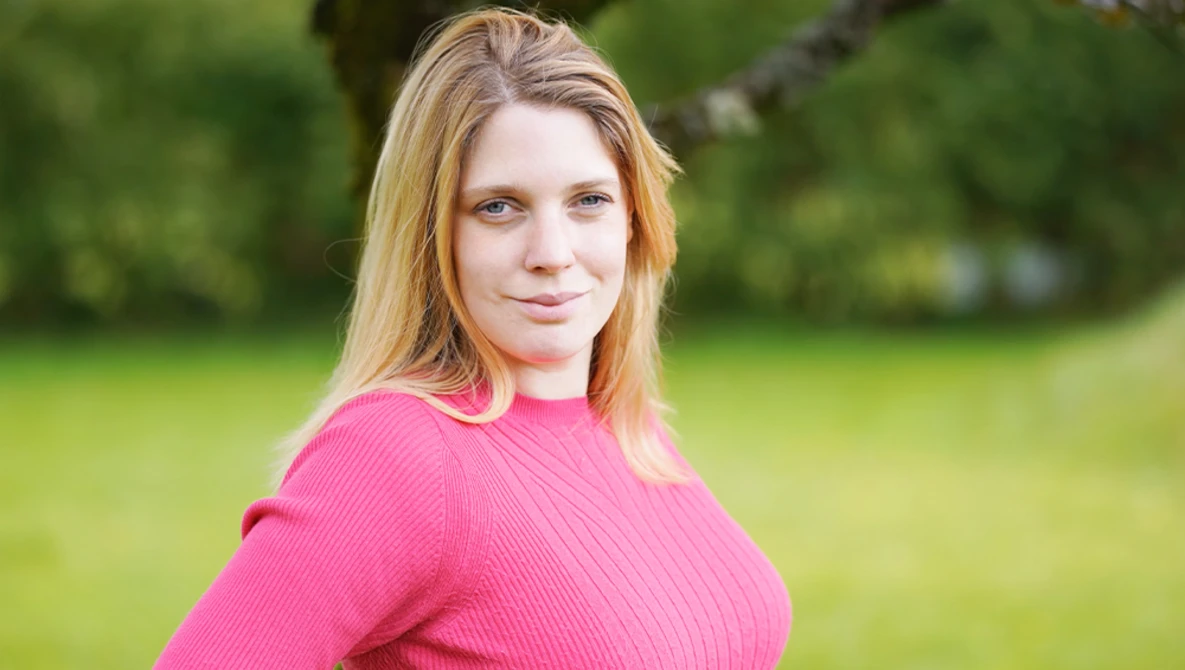 Woman in bright pink ribbed sweater poses outdoors against blurred green background.