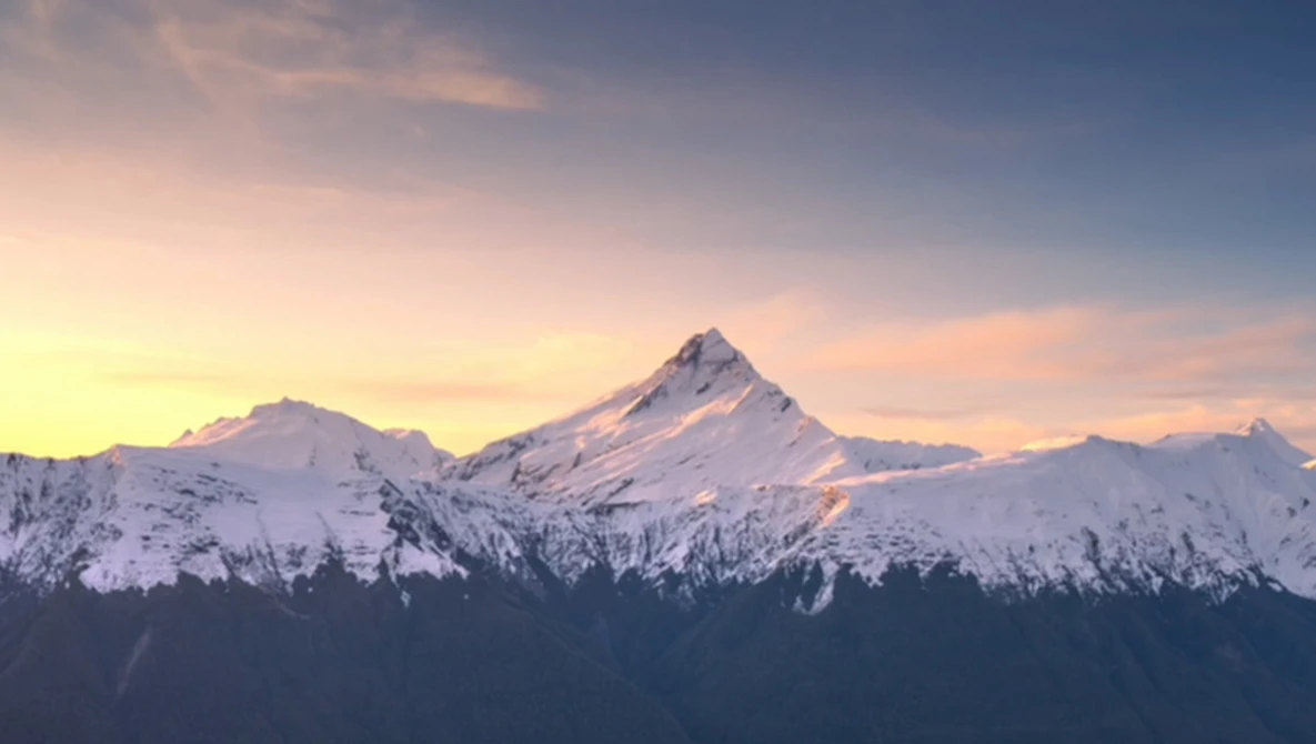 Snow-covered mountain peaks lit by golden sunrise light, with forested slopes in silhouette below.