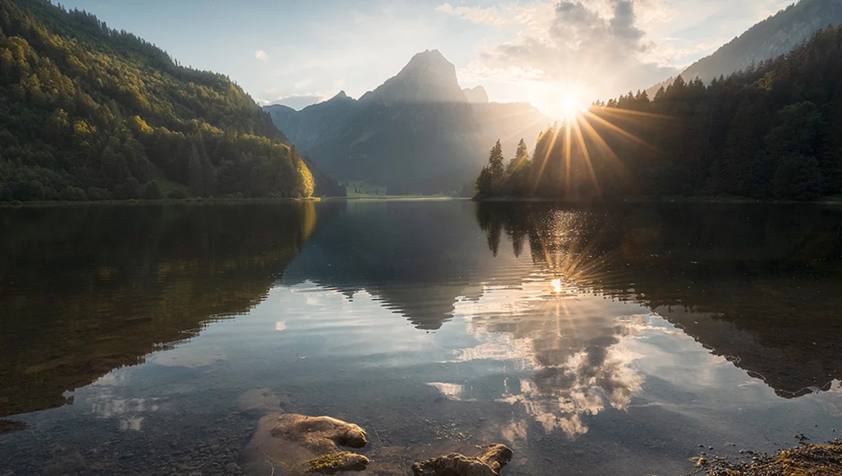 Mountain lake at sunrise with forested slopes and perfect mirror reflection in still water.