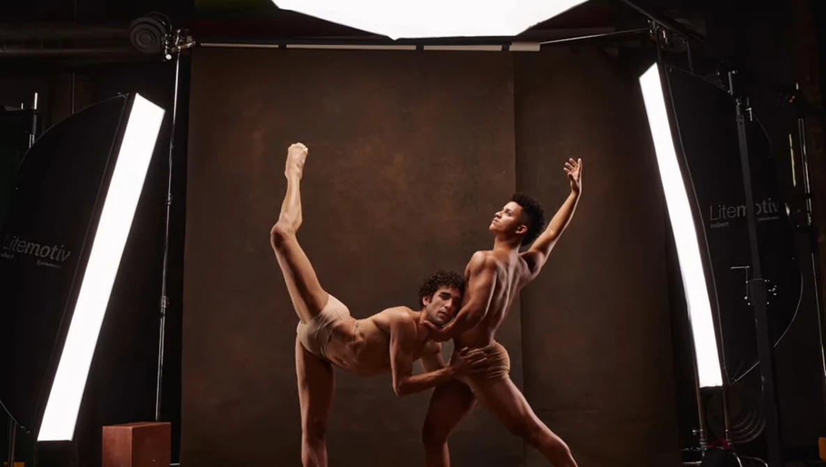 Two dancers in a studio performing a contemporary lift against a brown backdrop with professional lighting.