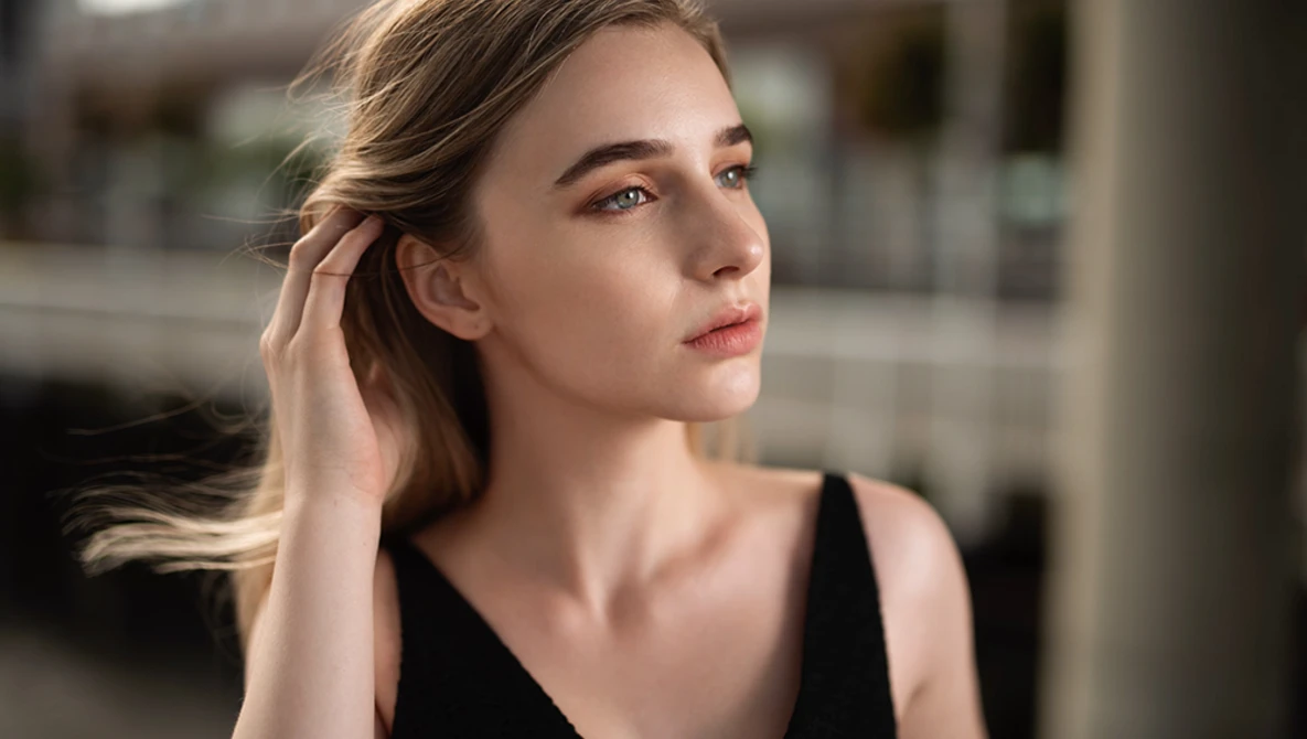Woman in black tank top looking away, hand near ear, soft natural lighting from the side.