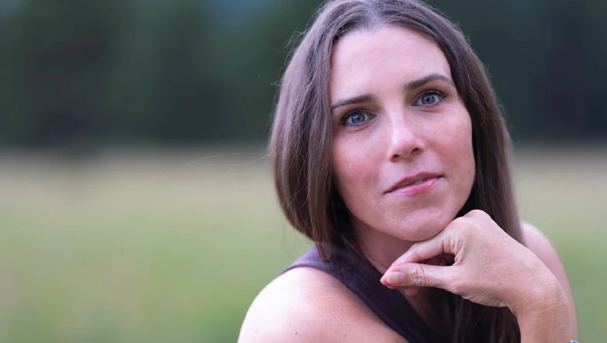 Woman with long brown hair looking over her shoulder outdoors with blurred green background.