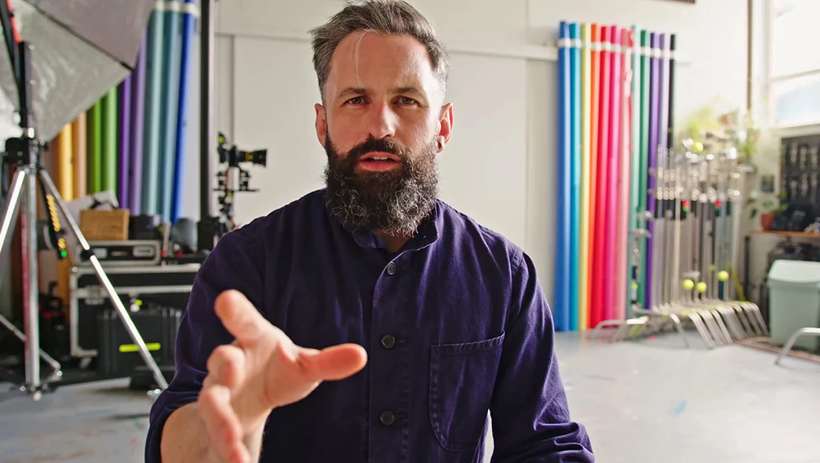 Bearded man in dark work shirt gesturing while speaking in a studio with colorful background rolls and lighting equipment.