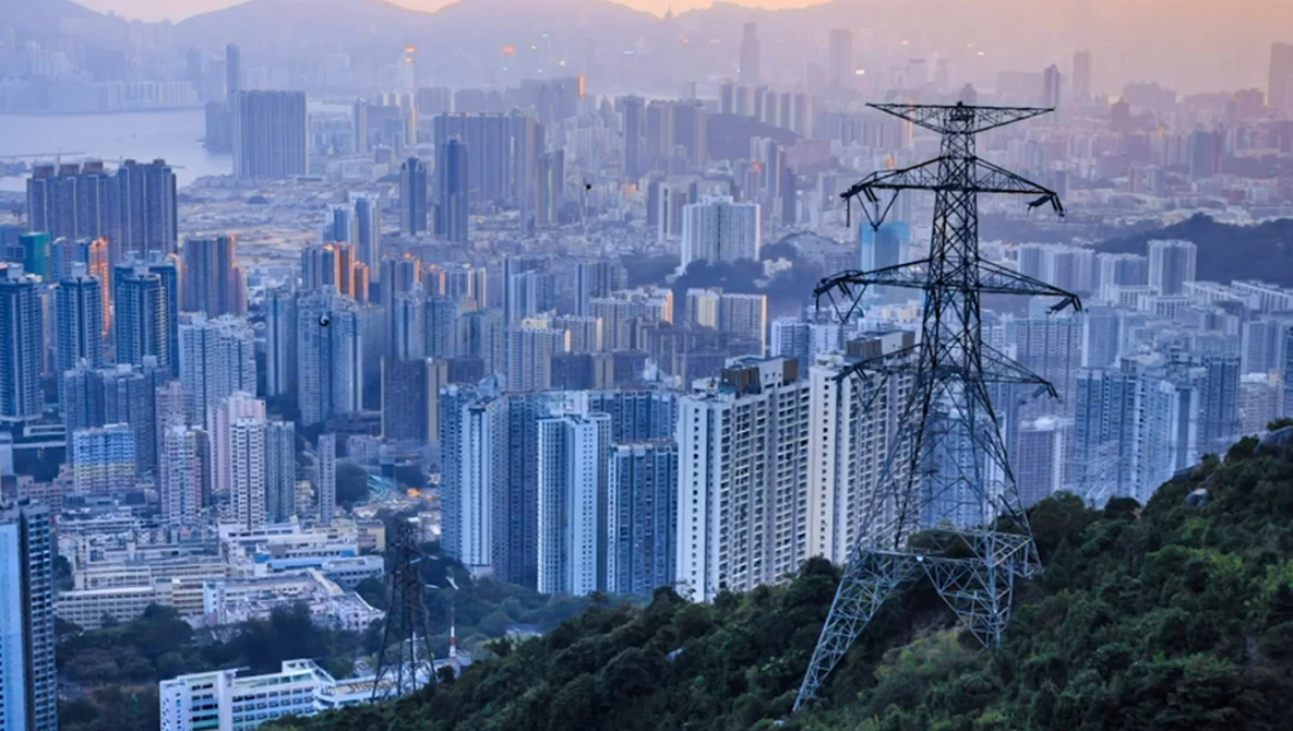 High-voltage transmission tower overlooking a sprawling cityscape at dusk with mountains in the distance.