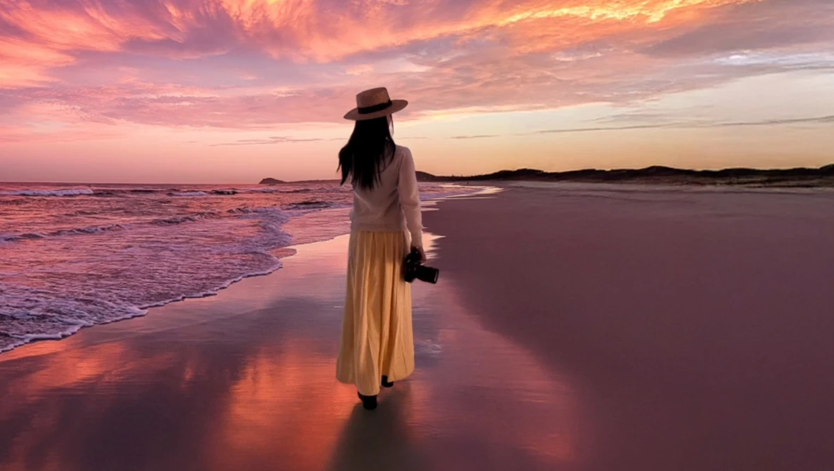 Woman in cream dress and hat standing in shallow ocean water during golden hour sunset.