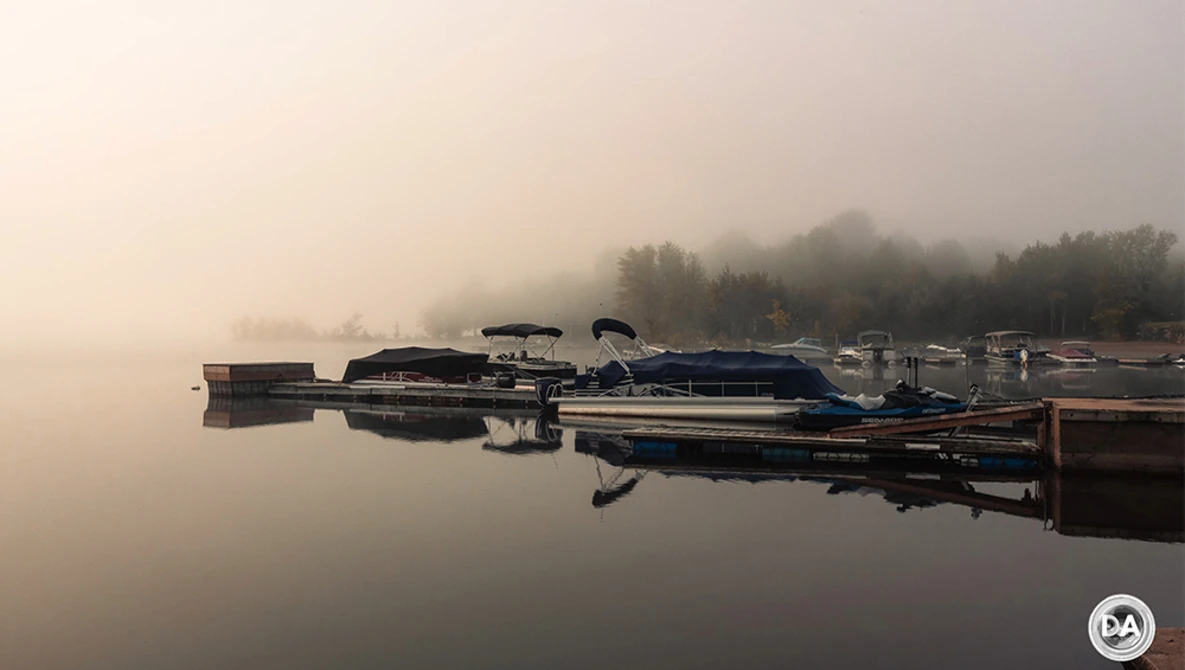 Moored boats reflected in still water at dawn with forested shoreline shrouded in fog.