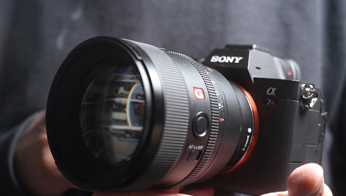Sony mirrorless camera with telephoto lens mounted, held in hands against blurred gray background.