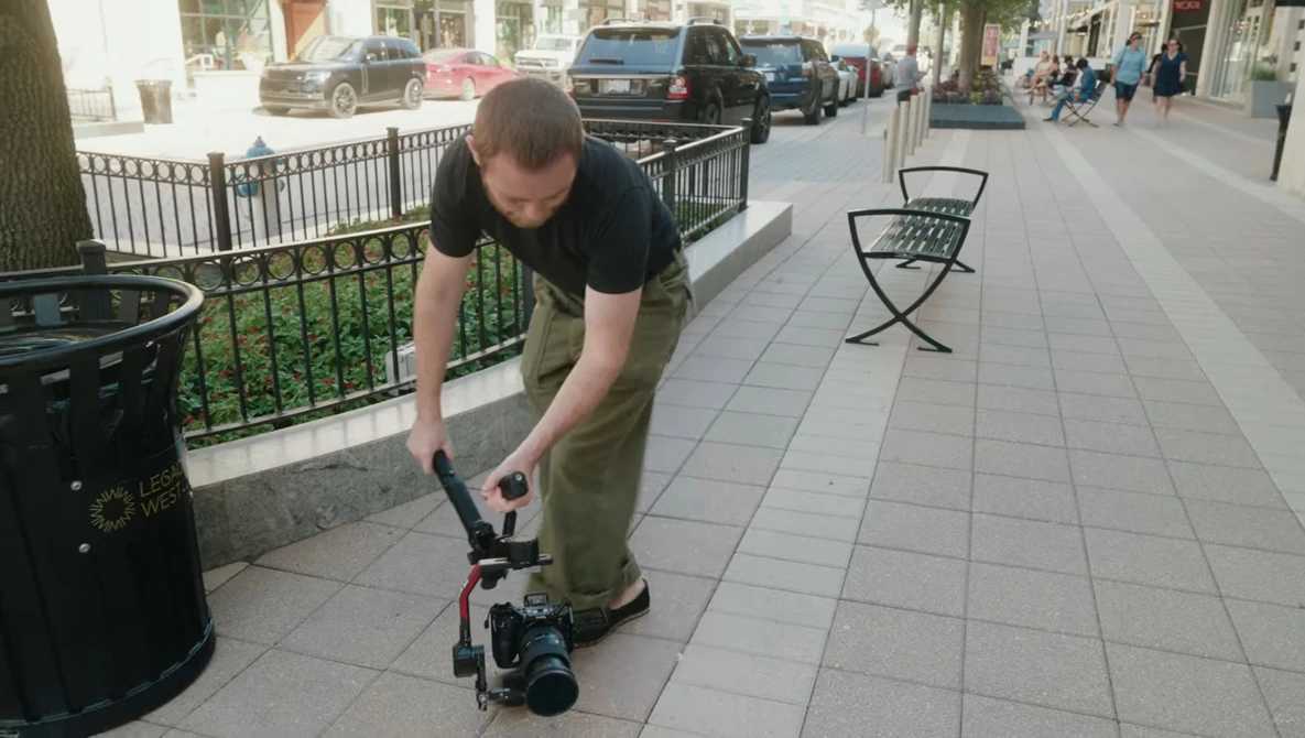 Photographer bending down to adjust a camera mounted on a skateboard on an urban sidewalk.