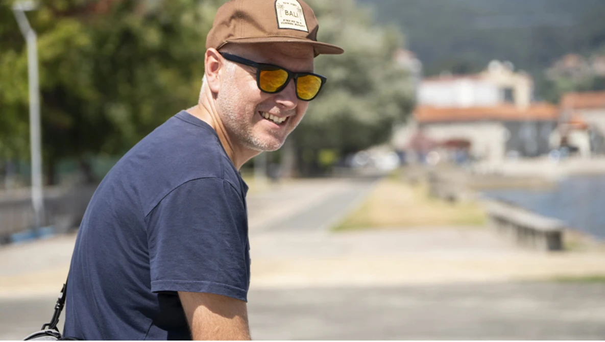 Man wearing a brown cap and yellow-tinted sunglasses smiles at the camera outdoors.