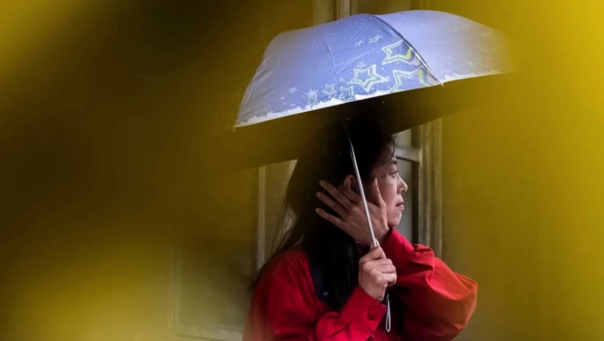 Woman in red jacket holding transparent umbrella against yellow background.