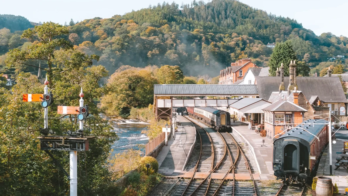 Steam locomotive stopped at a railway station beside a river with forested hills in autumn.