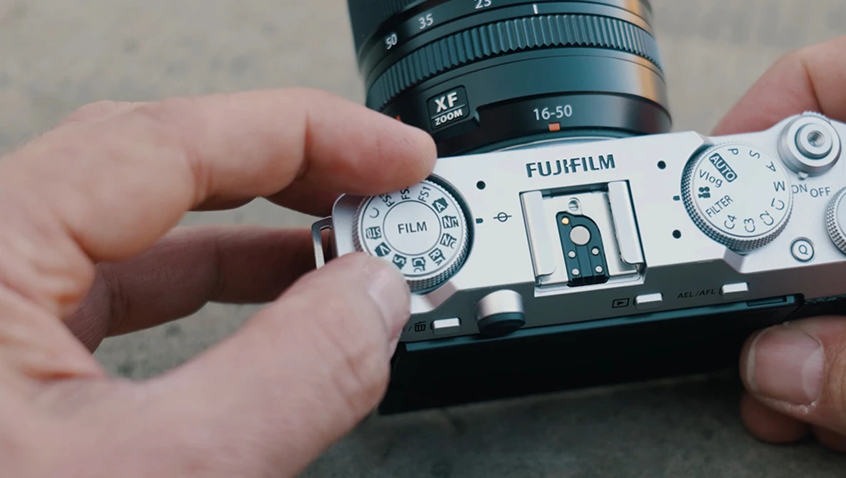 Hands holding a Fujifilm rangefinder camera with film advance dial visible and XF lens mounted on top.