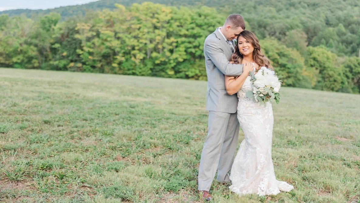 Bride and groom embracing in an open field with forested hills in the background.