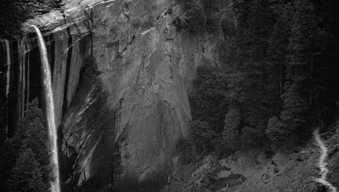 Waterfall cascading down a rocky cliff face surrounded by dense forest in black and white.