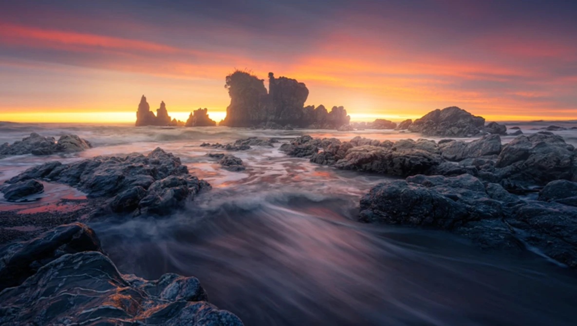 Dramatic coastal landscape with rocky outcrops and sea stacks silhouetted against a golden sunset.