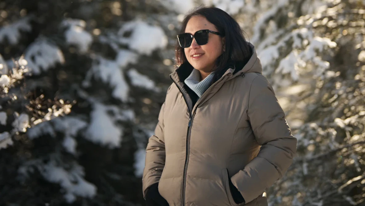 Woman in beige puffer jacket and sunglasses standing in snowy forest with evergreen trees.