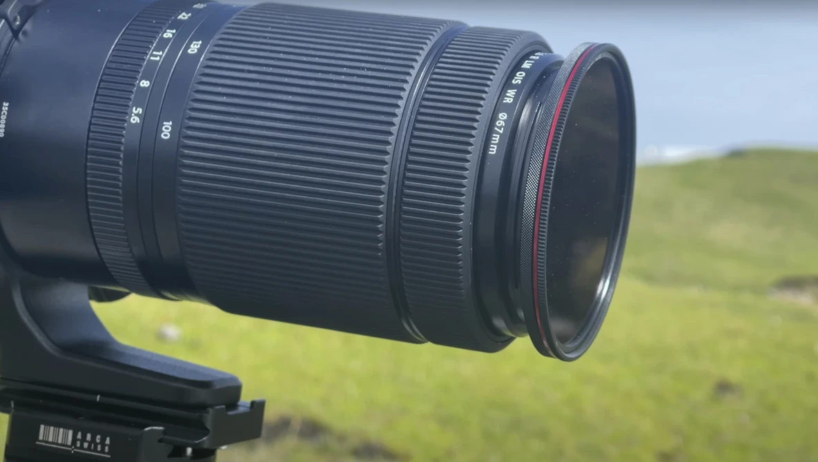 Telephoto zoom lens mounted on camera body with grassy hillside in soft focus background.