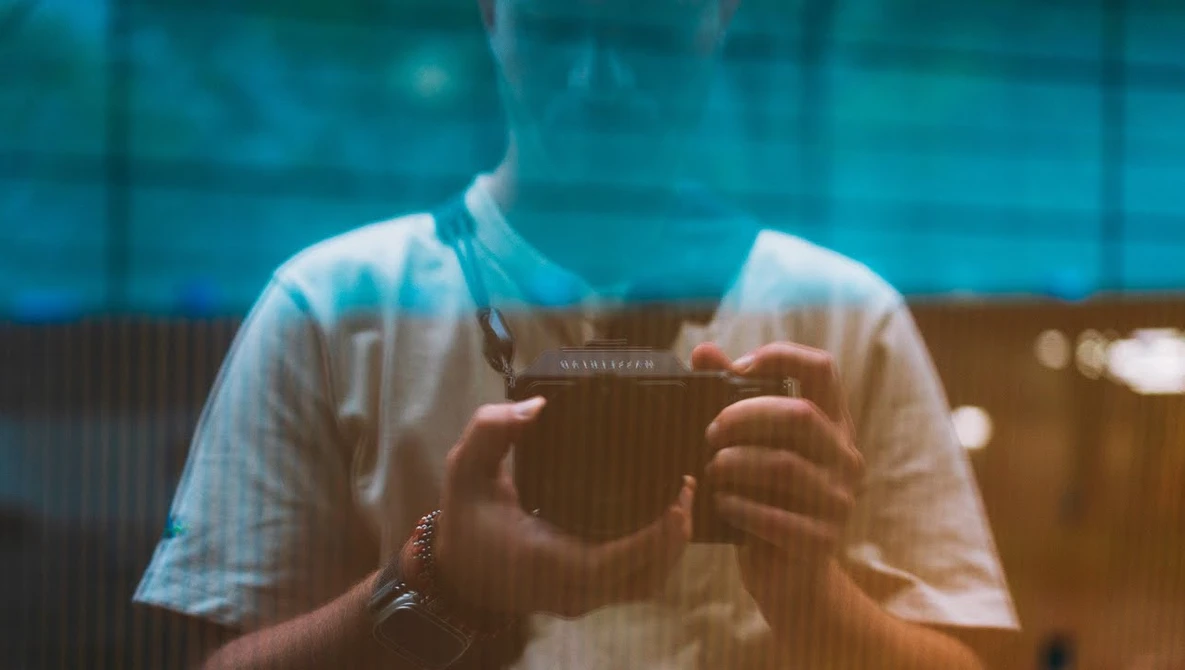 Photographer holding a vintage camera with blue-tinted window reflections obscuring their face.