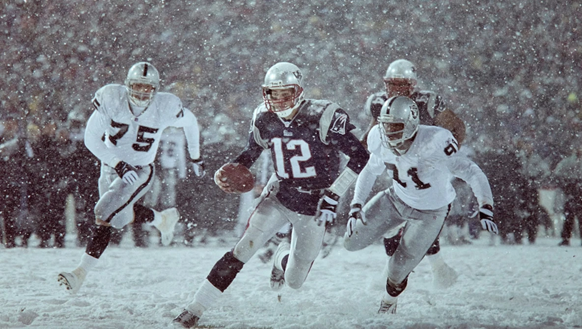 Four football players in a heavy snowstorm, two wearing white uniforms and two in dark uniforms with numbers visible.