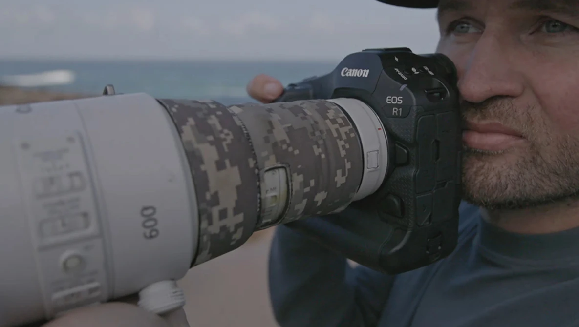 Photographer holding Canon EOS R1 with white telephoto lens featuring digital camouflage pattern.