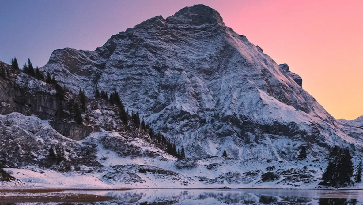 Snow-covered mountain peak reflected in a partially frozen lake at sunset, with evergreen trees along the shoreline.