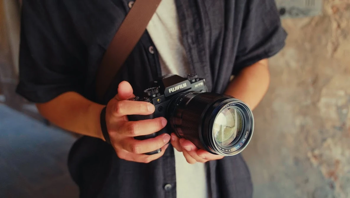 Photographer holding a black Fujifilm camera with a telephoto lens against their chest.