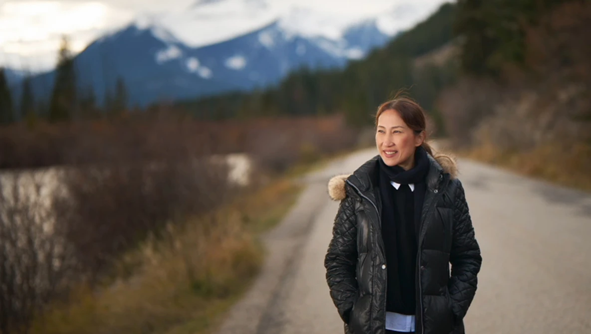 Woman in black quilted jacket smiling at camera on rural road with snow-capped mountains and river in background.