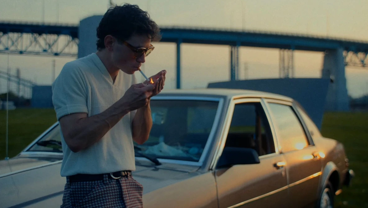 Man in glasses and white shirt eating a slice of pizza outdoors beside a vintage sedan.